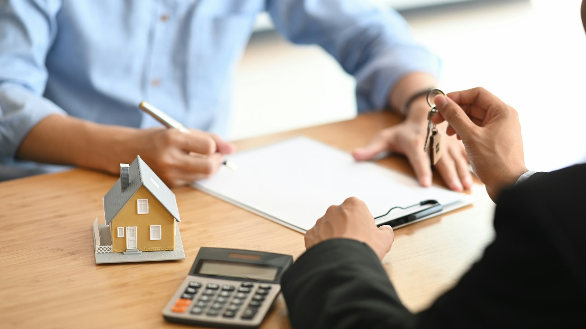 Mortgage broker handing house keys to client during a consultation with model home and calculator on desk